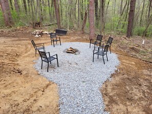 A circular seating area with four chairs around a central fire pit, surrounded by gravel and dirt, set in a forested area. - Jack of Hearts. Secluded, Hocking Hills cabin! (Rockbridge)