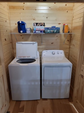 A compact laundry room with two front-loading washing machines and a shelf above containing board games and a bottle. - Jack of Hearts. Secluded, Hocking Hills cabin! (Rockbridge)