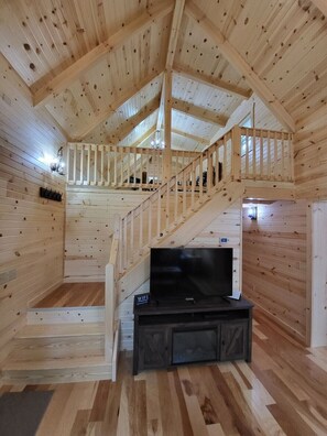 A wooden cabin interior with a TV, wooden floors, a staircase, and a balcony.