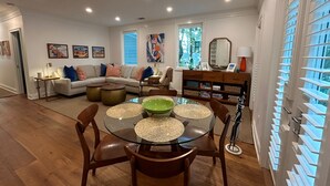Un salon moderne avec une table à manger en verre, des chaises en bois, un canapé et une table basse. On aperçoit une fenêtre avec des stores et une bibliothèque au fond.