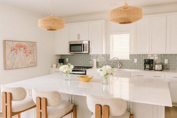 A modern kitchen with white cabinetry, a central island with chairs, a microwave, and a coffee maker. - Coastal Home · Bay Views · Near the Wharf · Pet Friendly (Orange Beach)