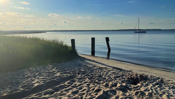 Hiekkainen ranta, tyyni meri, kauempana näkyvä purjevene ja kirkas taivas.
