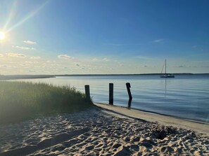 Ein sandiger Strand mit ruhiger See, ein Segelboot in der Ferne und ein klarer Himmel.