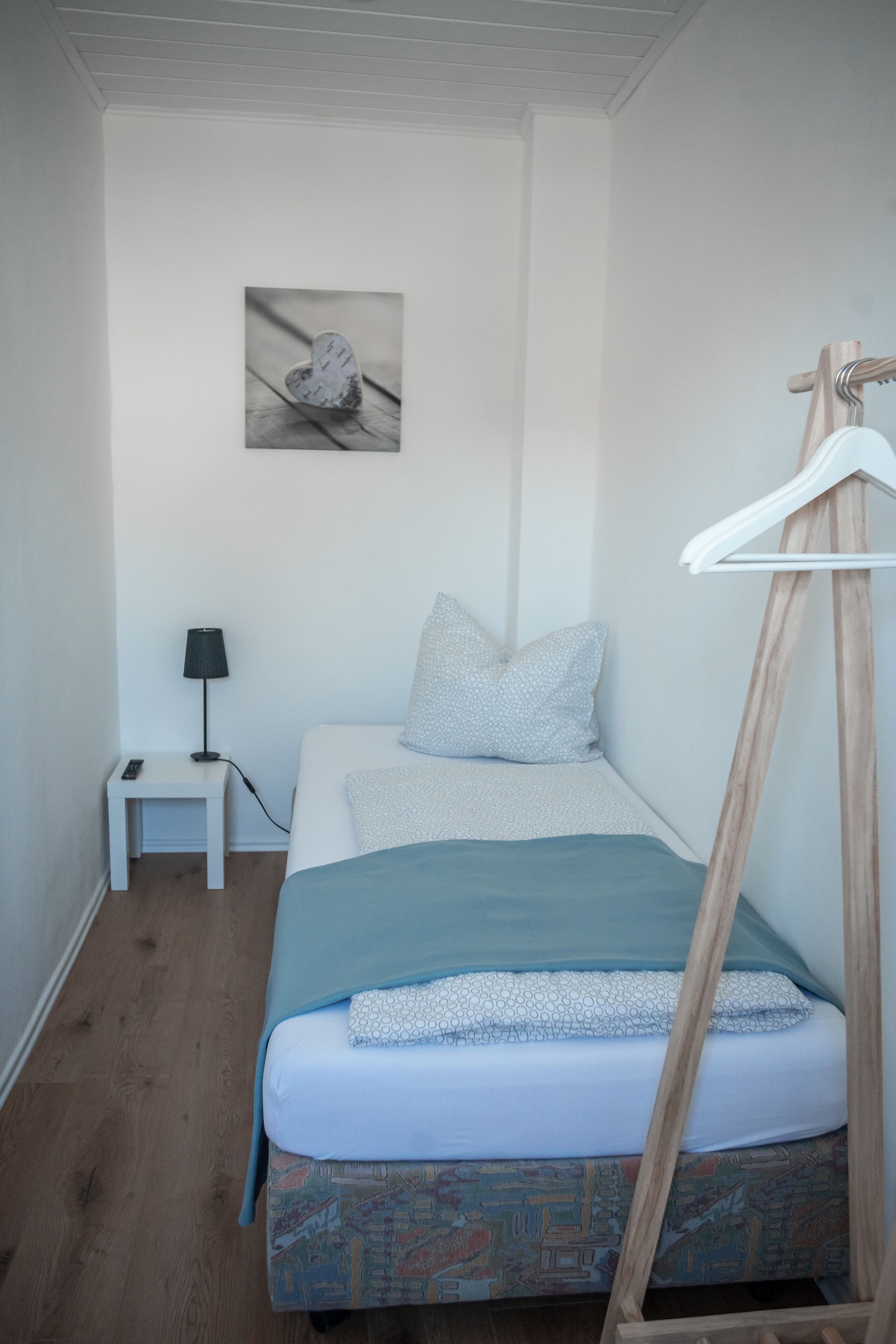 A small, neatly arranged bedroom with a single bed, a bedside table with a lamp, and a wall-mounted picture.