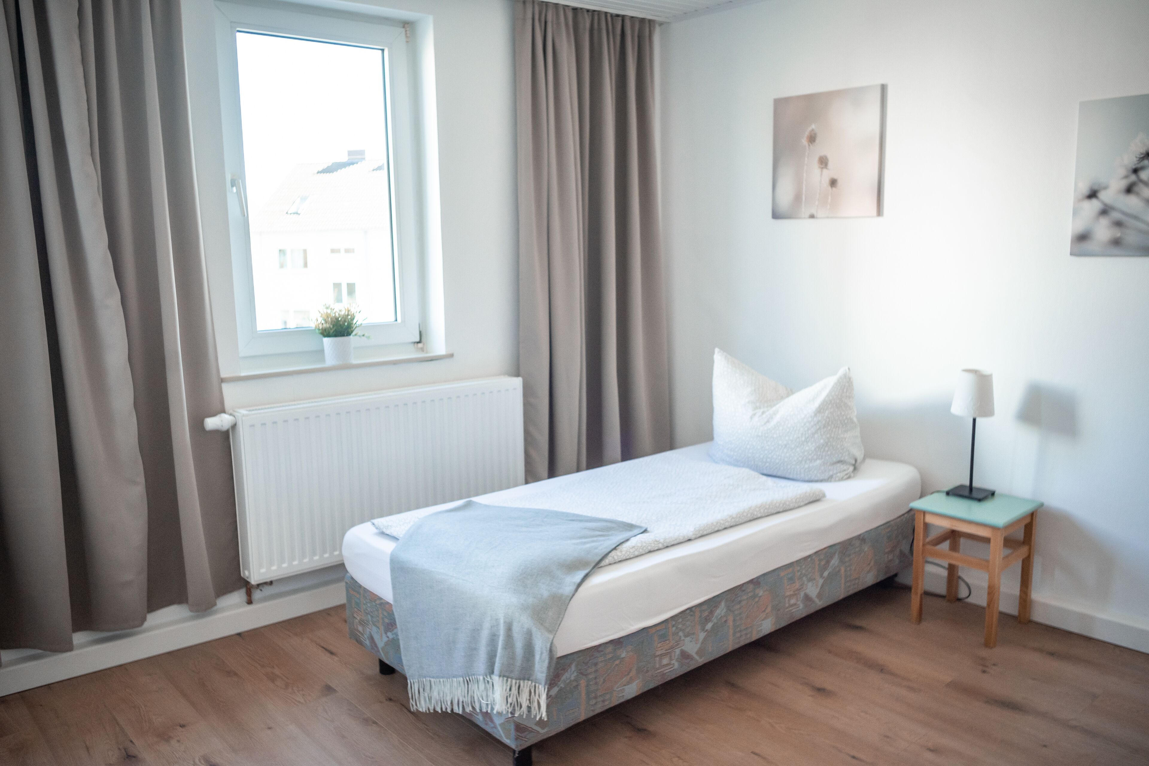 Ein Zimmer mit einem Bett, einem Vorhangfenster, einer Heizung, einem kleinen Tisch mit Lampe und einem Bild an der Wand.