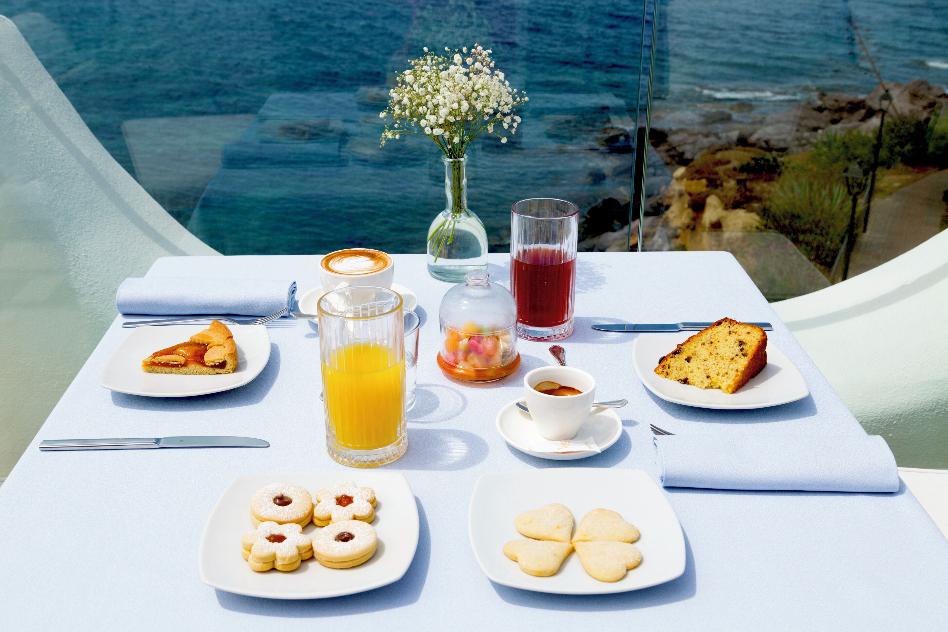 A table set with breakfast items including pastries, a glass of orange juice, and a vase of flowers, overlooking a body of water.