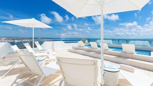 A rooftop pool area with white lounge chairs and umbrellas, overlooking a cityscape and the sea. - Relais la Ghinghetta (Portoscuso)