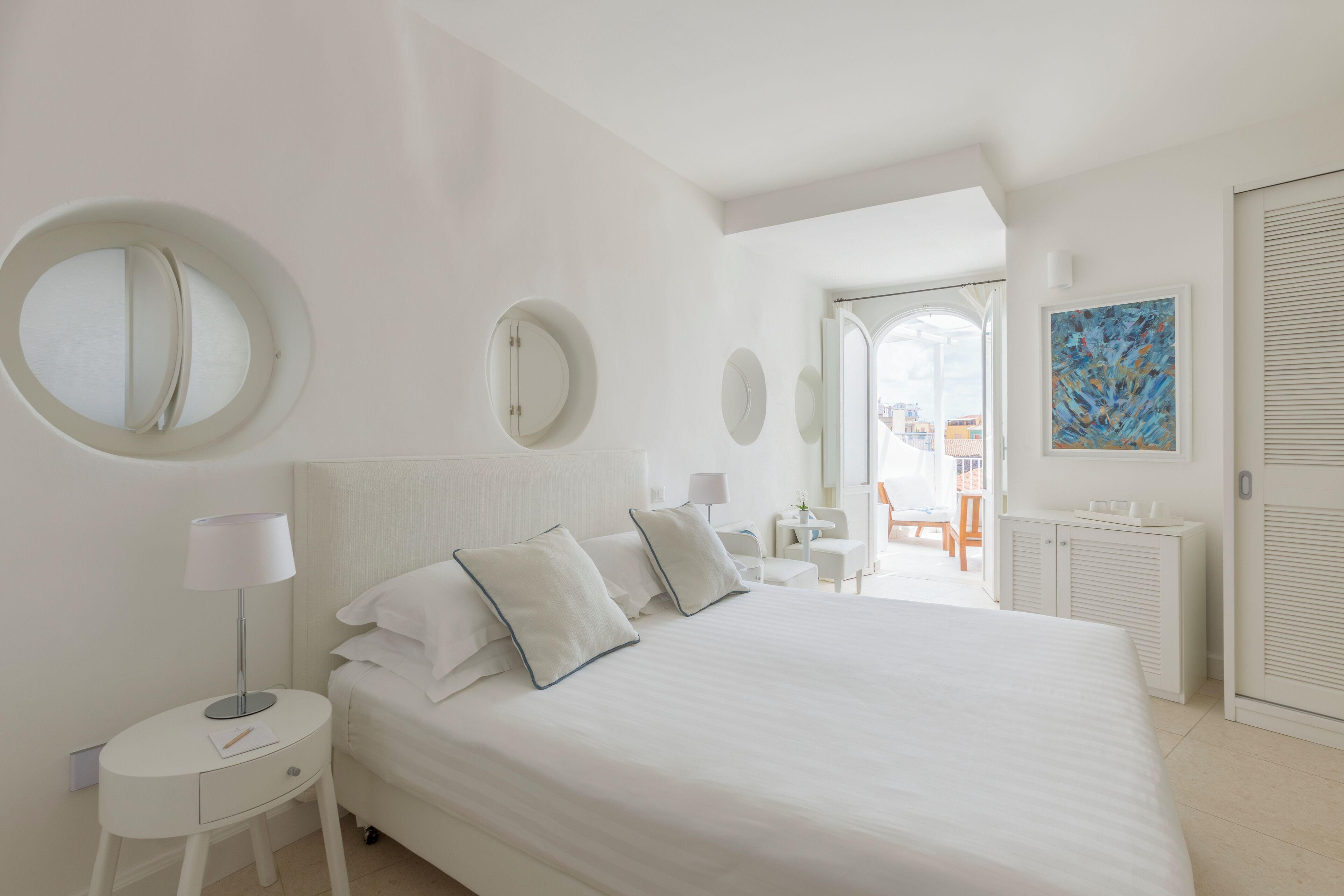 A modern hotel room with a large bed, bedside tables, a round mirror, a painting, and a view of the outside.