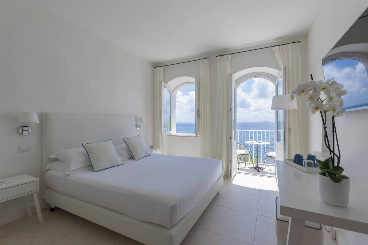 A white bedroom with a bed, a nightstand, a desk, a potted orchid, and a balcony with a view of the sea.
