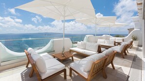 A sunlit outdoor seating area with white cushioned sofas and wooden chairs, large white umbrellas, and a view of the sea. - Relais la Ghinghetta (Portoscuso)