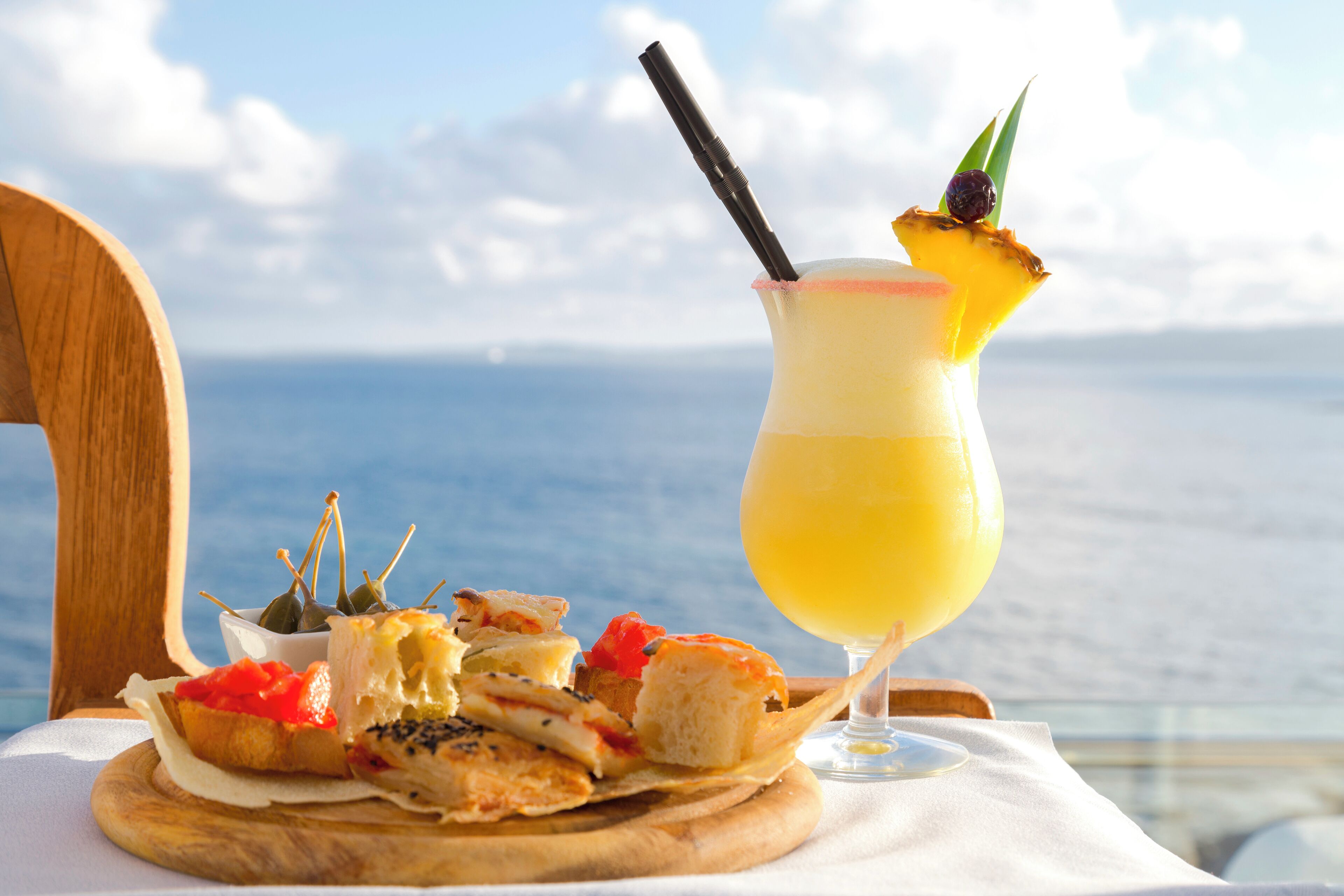 A wooden table with a variety of appetizers and a glass of a yellow beverage with a straw and a garnish, set against a scenic ocean view.