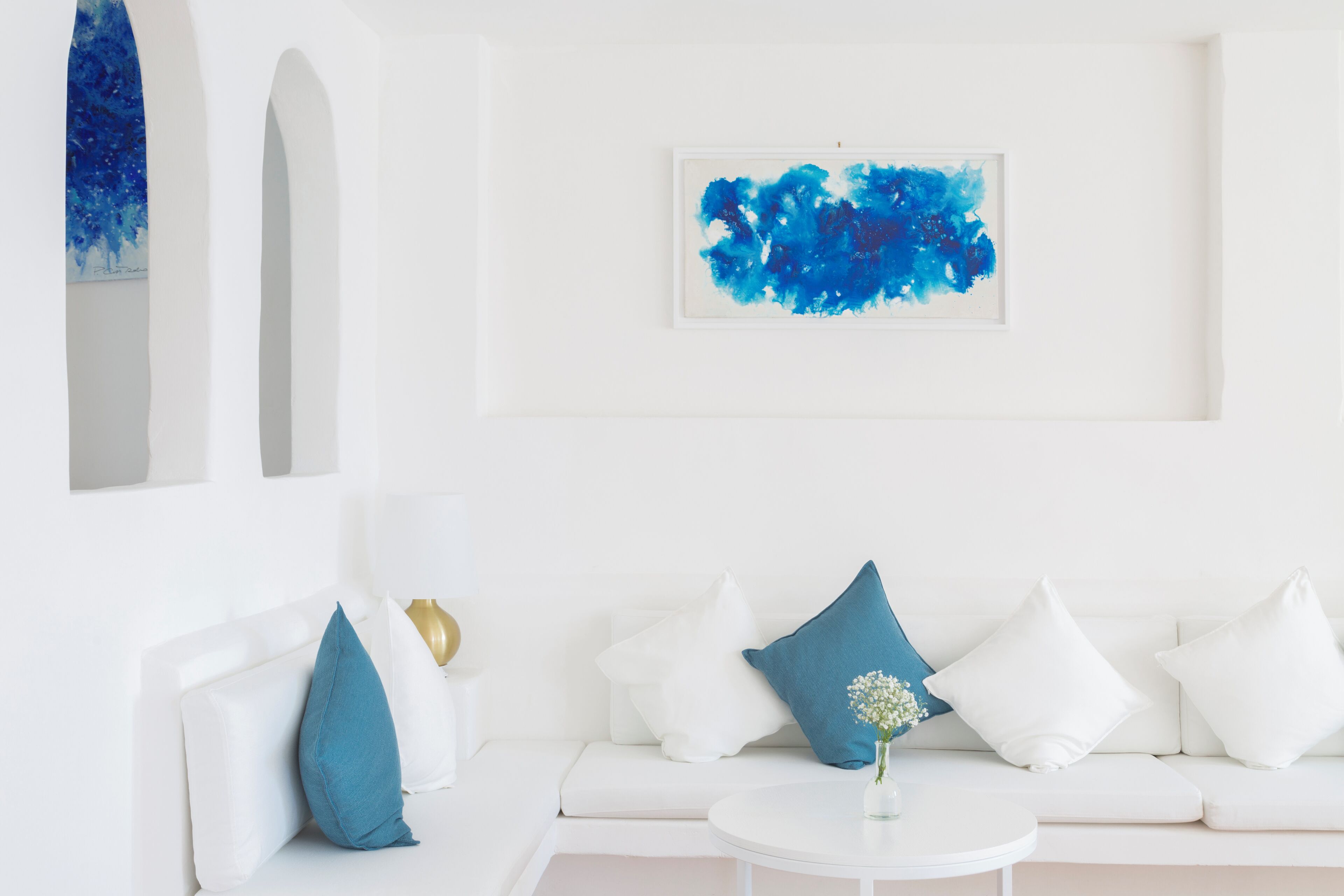 A modern living room with a white sofa, blue and white cushions, a round coffee table, and a blue abstract painting on the wall.