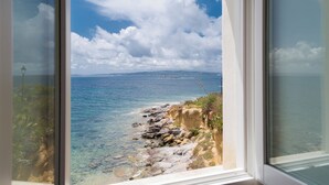 A coastal view through a window, featuring clear blue water, rocky shoreline, and distant land. - Relais la Ghinghetta (Portoscuso)
