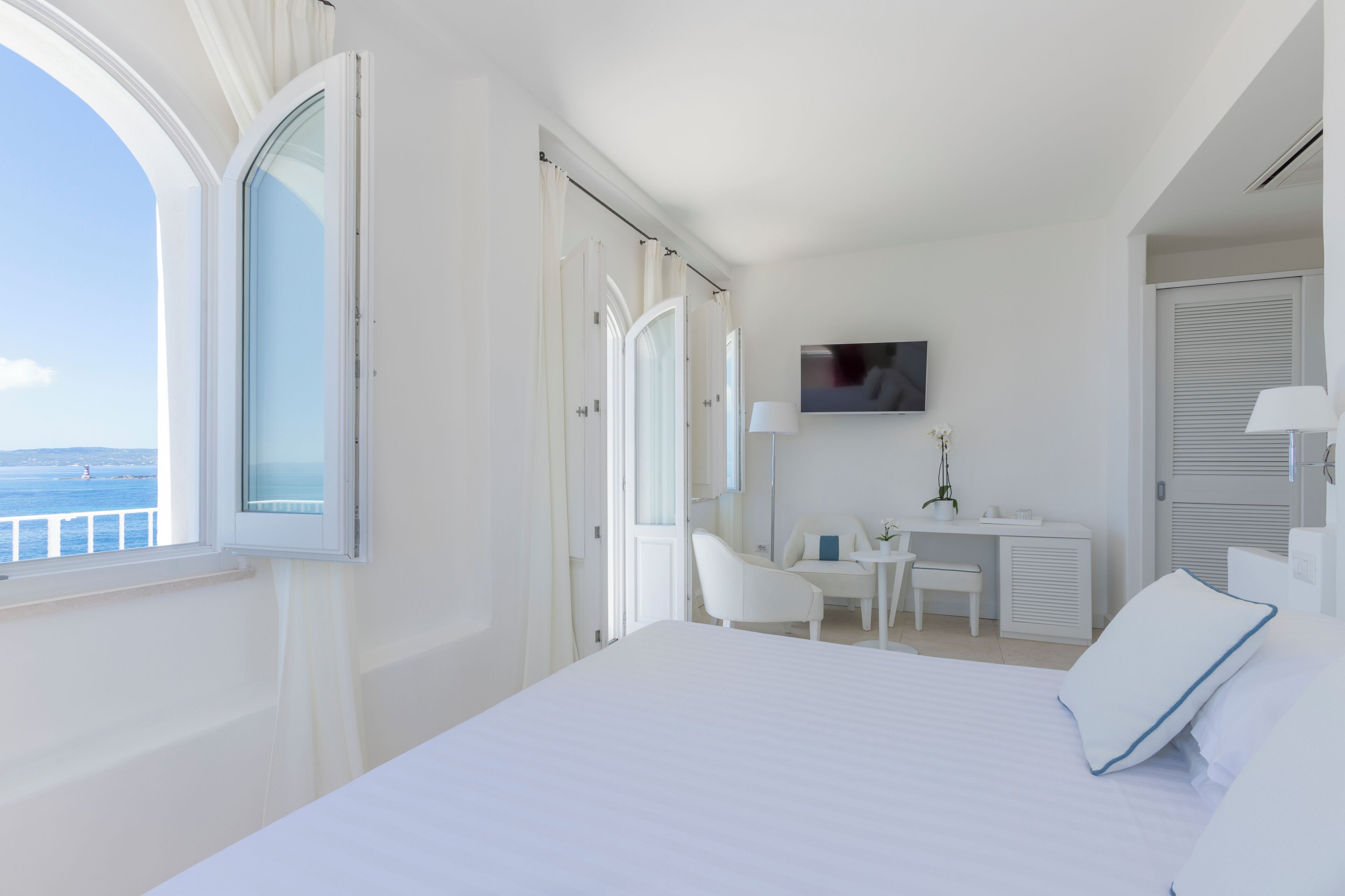 A hotel room with a bed, a desk, a chair, a TV, and a view of the sea.