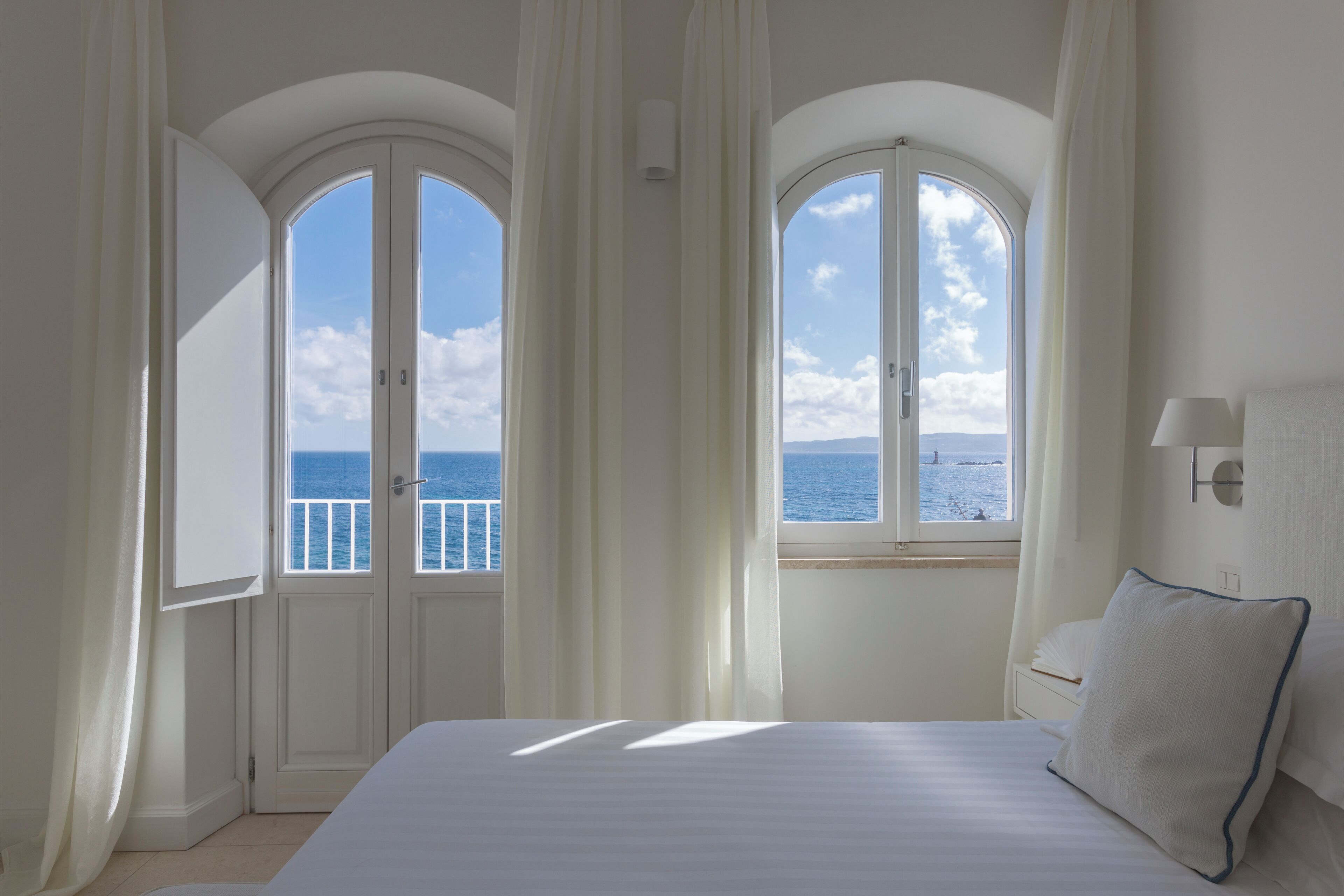 A bedroom with a large bed, two windows with curtains, a view of the sea, and a nightstand with a lamp.