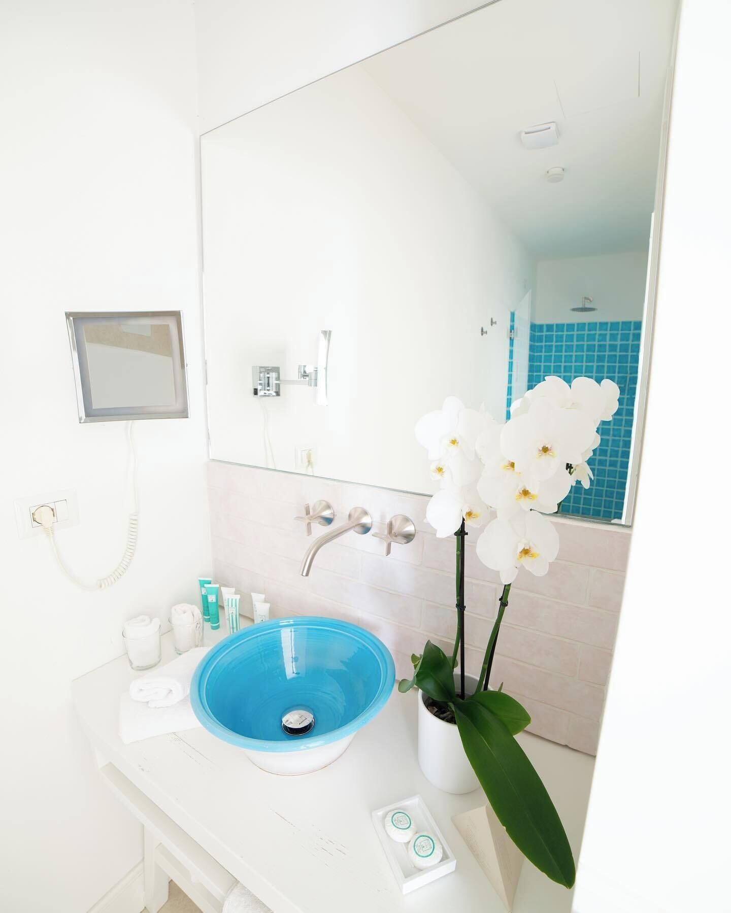 Un baño con un lavabo blanco, una pileta azul y un espejo grande. Hay una planta de orquídea blanca en la encimera y una pared de ducha con azulejos azules al fondo.