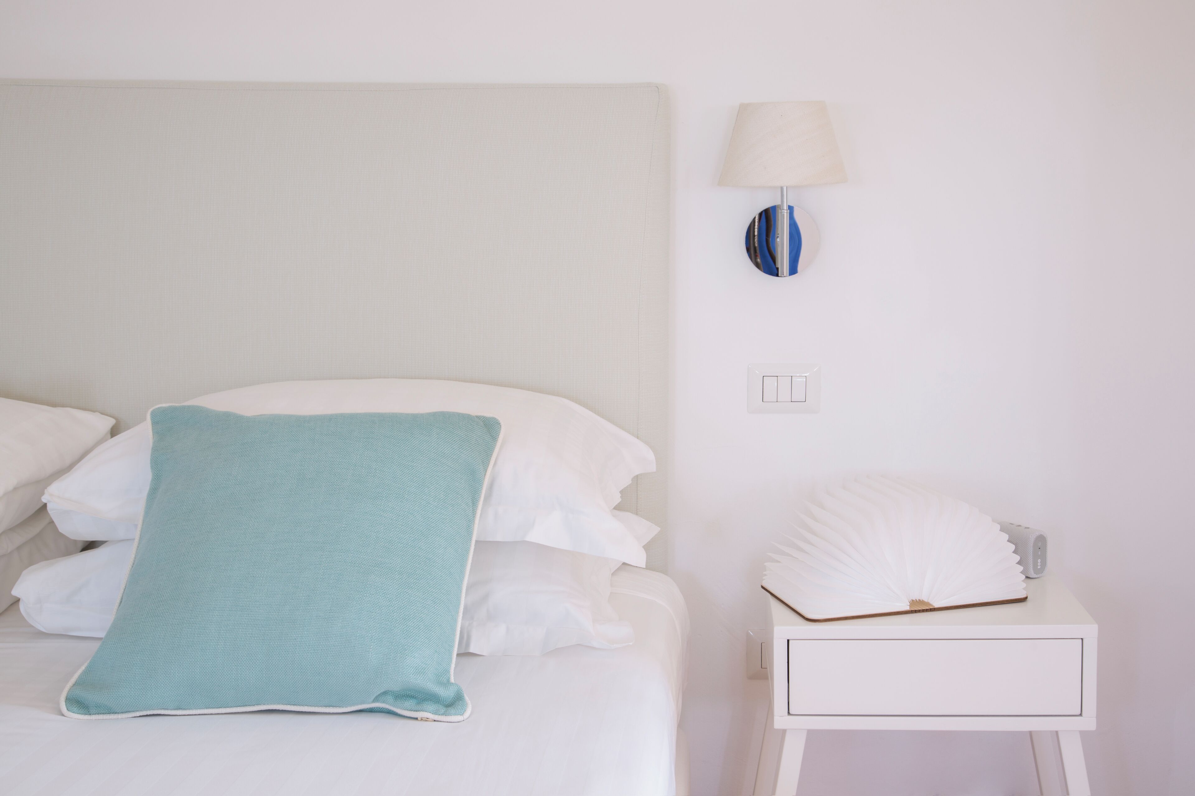 A neatly made bed with white and teal pillows, a white nightstand with a lamp, and a decorative shell on the nightstand.