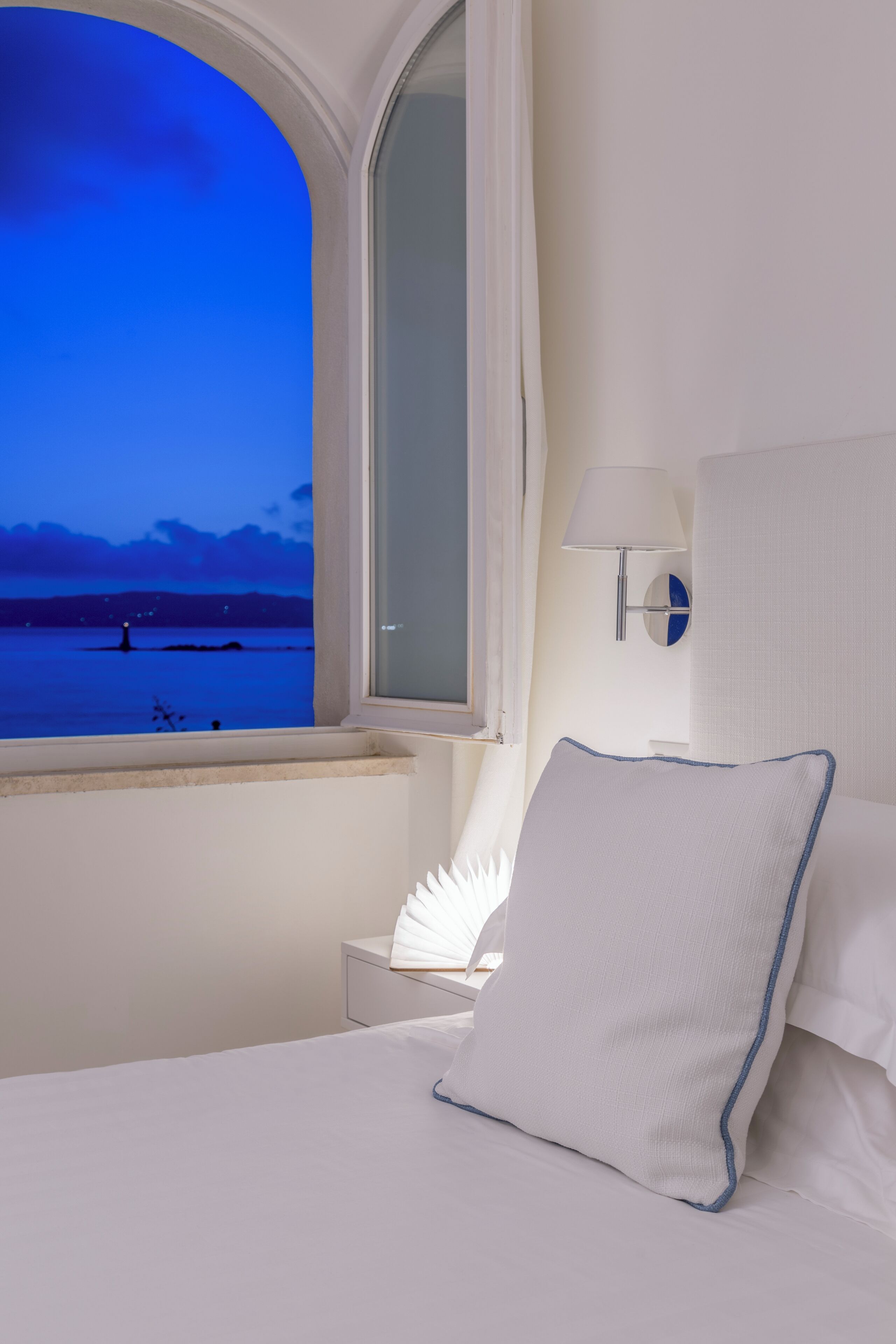 Ein Hotelzimmer mit Bett, Nachttisch, Lampe, Spiegel und Fenster mit Blick auf ein Gewässer im Abenddämmerung.
