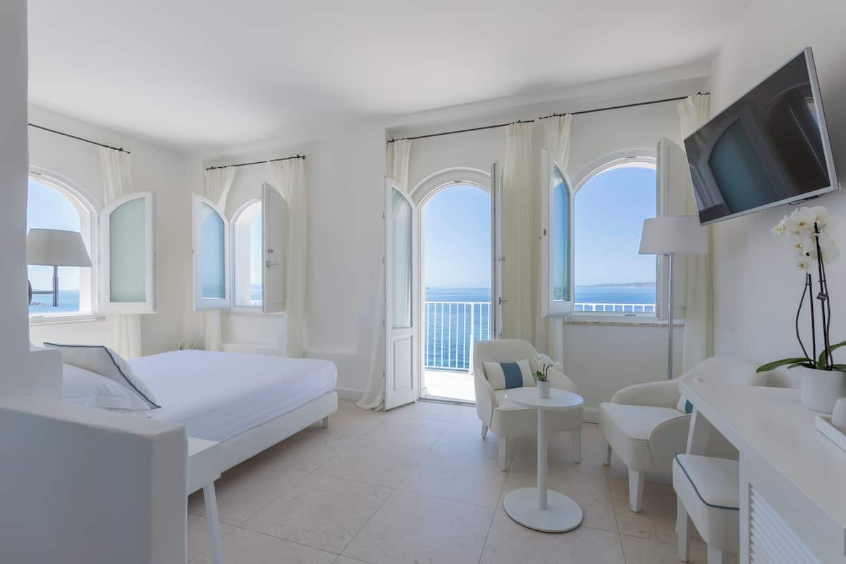 A hotel room with a bed, a desk, a chair, a TV, and a balcony with a view of the sea.