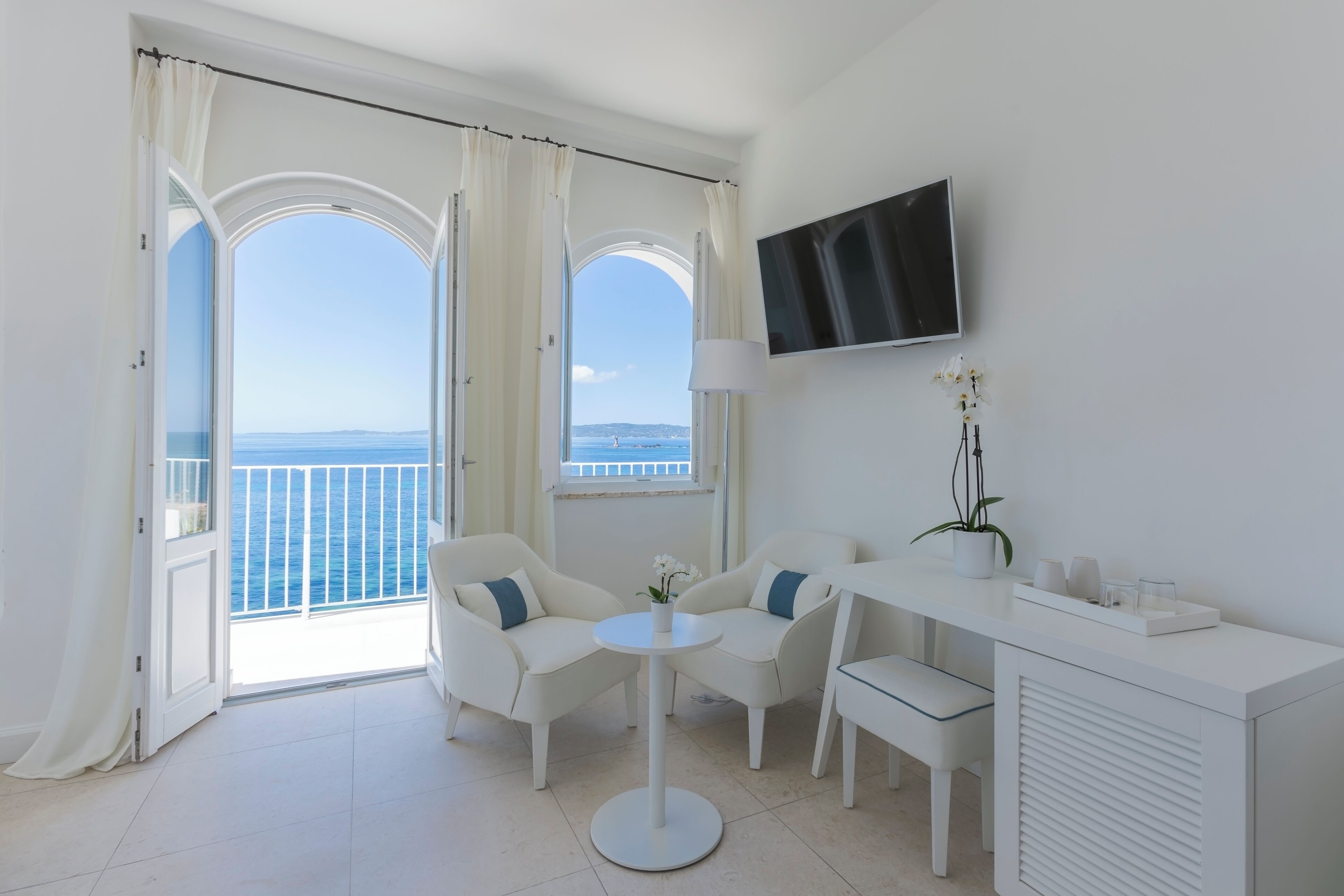 Una habitación con balcón, muebles blancos, televisor y una planta.