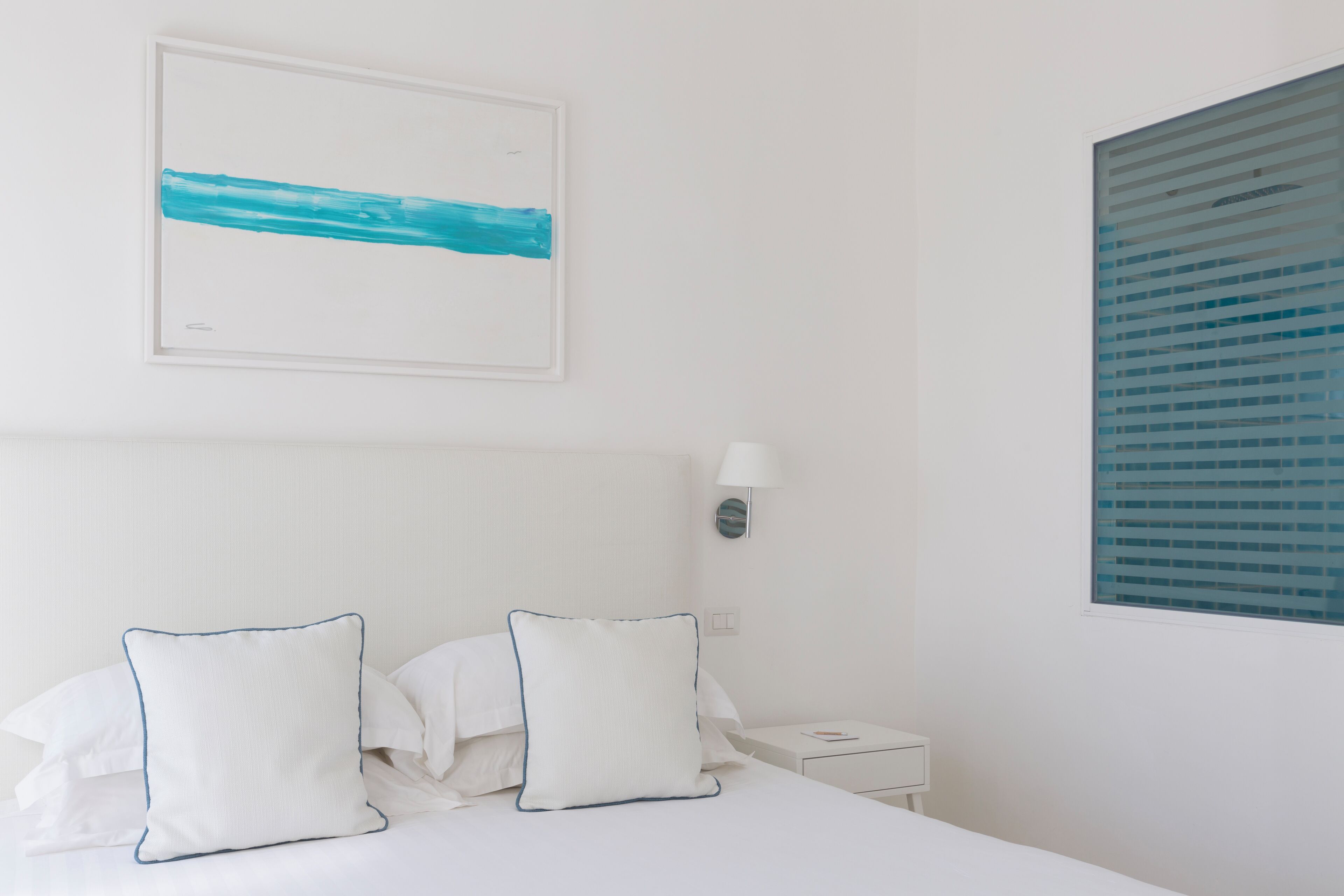 A neatly made bed with white linens and two pillows, a bedside table with a lamp, and a framed abstract painting on the wall.