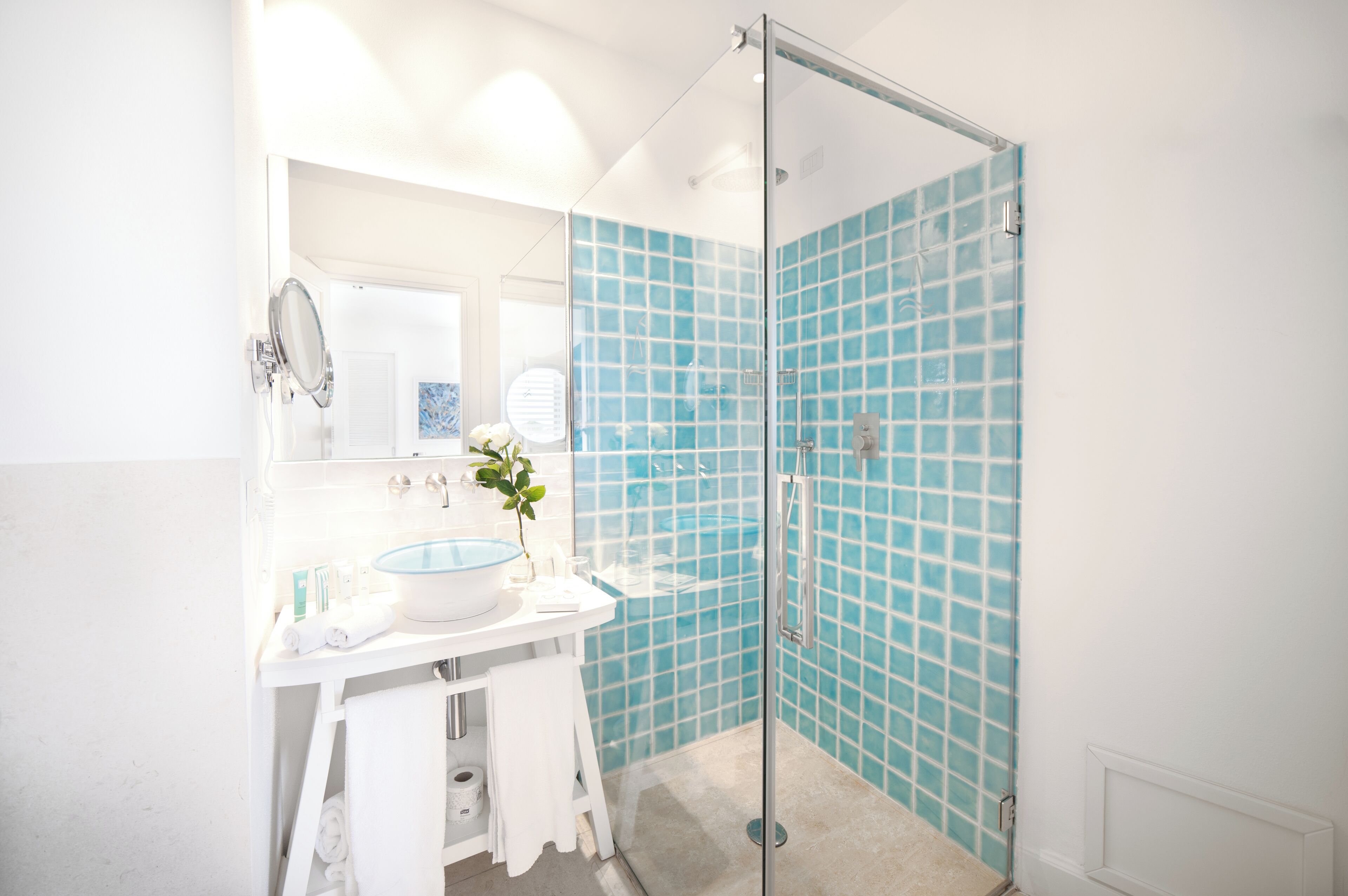 Un baño con ducha envuelta en vidrio, un lavabo blanco con recipiente azul y un espejo sobre el lavabo.