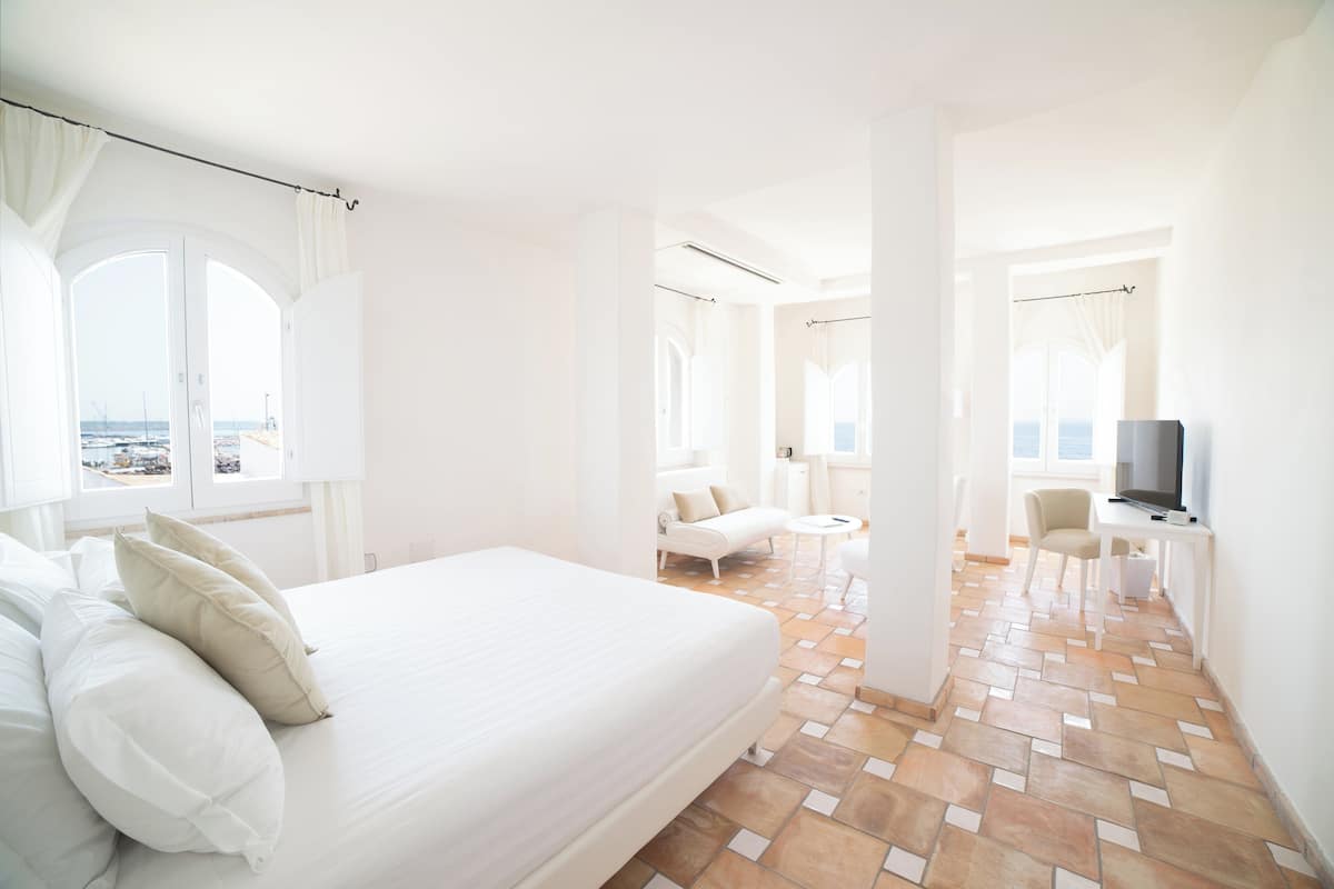 A hotel room with a large bed, a TV, a desk, and a view of the sea.