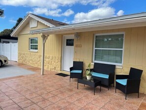 A yellow building with a stone facade, a white door, and a large window. There are two black chairs with blue cushions and a small table with a potted plant.