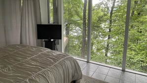 Camera da letto con un ampio letto, una TV a schermo piatto e una porta scorrevole in vetro che dà su un balcone con vista sulla foresta.