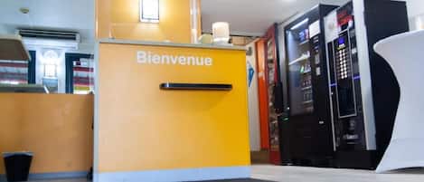 A reception area with a 'Bienvenue' sign, a black trash bin, and vending machines.