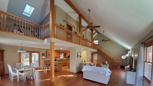 A spacious loft-style living area with high ceilings, wooden floors, a dining area with white furniture, and a staircase with a wooden railing.