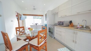 A modern kitchen with white cabinets, a stainless steel oven, and a microwave. There is a dining table with chairs and a fruit bowl on it.