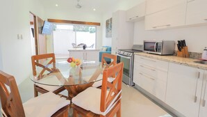 A modern kitchen with white cabinets, a built-in oven, a microwave, and a sink. There is a dining area with a glass table and wooden chairs.