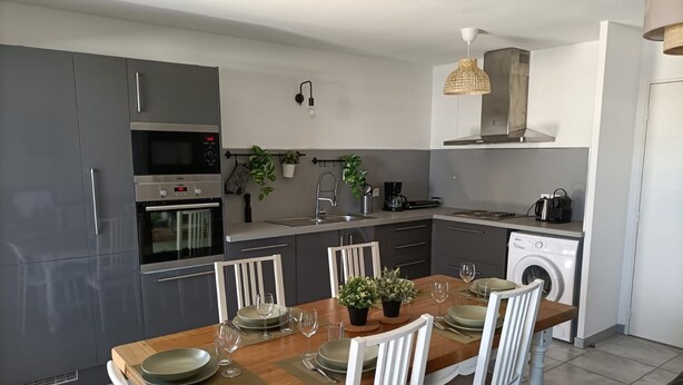 Eine moderne Küche mit grauen Schränken, einem eingebauten Backofen, einer Mikrowelle und einer Waschmaschine. Ein Esstisch mit weißen Stühlen und einer Holzplatte steht dort.