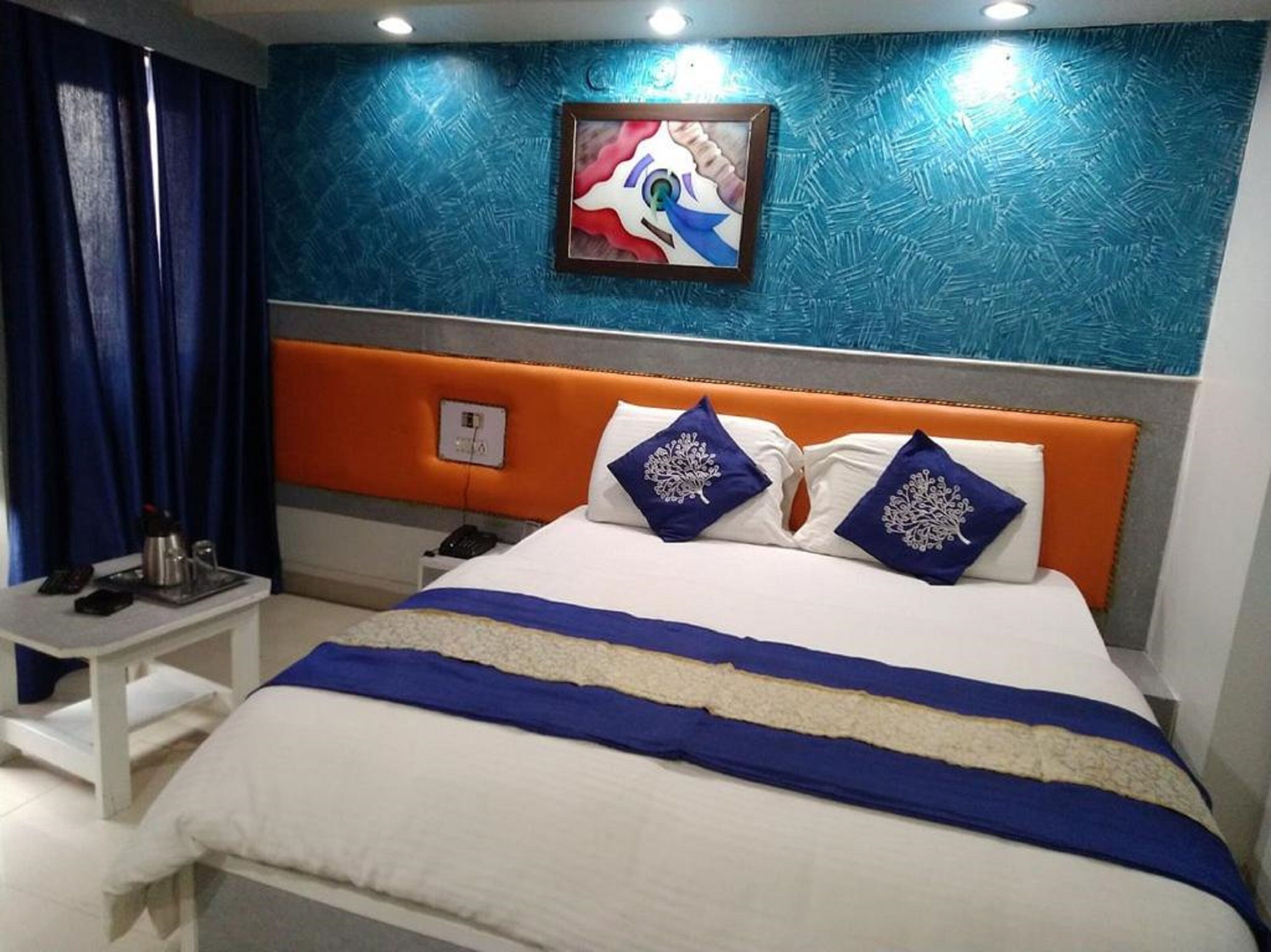 A modern hotel room with a large bed, blue and white bedding, a decorative pillow, a bedside table, and a painting on the wall.
