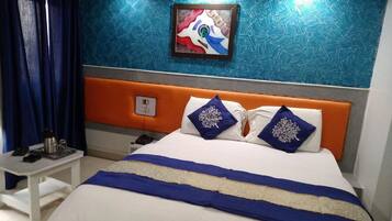 A modern hotel room with a large bed, blue and white bedding, a decorative pillow, a bedside table, and a painting on the wall.