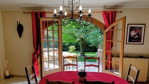 Ein Essbereich mit einem runden Tisch, der mit einer roten Tischdecke bedeckt ist, umgeben von Holzsesseln. Über dem Tisch hängt eine Chandeliebre, und ein Fenster mit roten Vorhängen gibt auf einen Garten hinaus.