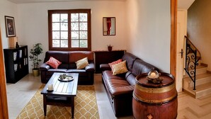 Ein Wohnzimmer mit braunem Ledersofa, einem Holztisch, einem gemusterten Teppich, einem Bücherregal, einem Blumentopf und einem Holzfässchen.