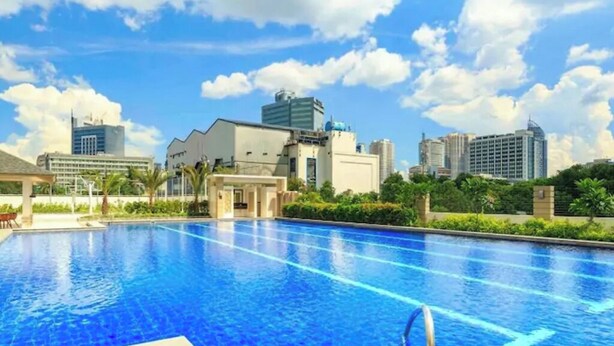 A swimming pool with clear blue water, surrounded by greenery and modern buildings in the background.