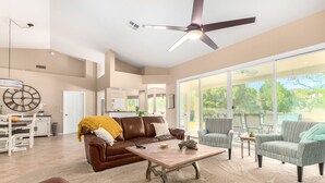 Ein geräumiges Wohnzimmer mit einem braunen Ledersofa, einem Couchtisch und gestreiften Sesseln. Ein Deckenventilator und große Fenster sorgen für natürliches Licht.