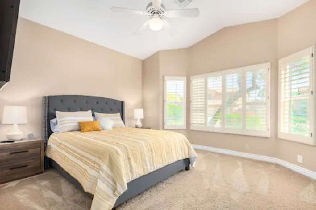 A bedroom with a bed, nightstands, a ceiling fan, and large windows with blinds.