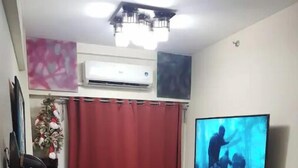 A living room with a black leather sofa, a red curtain, a television, and an air conditioning unit. - Mucucu's place (Pasig)