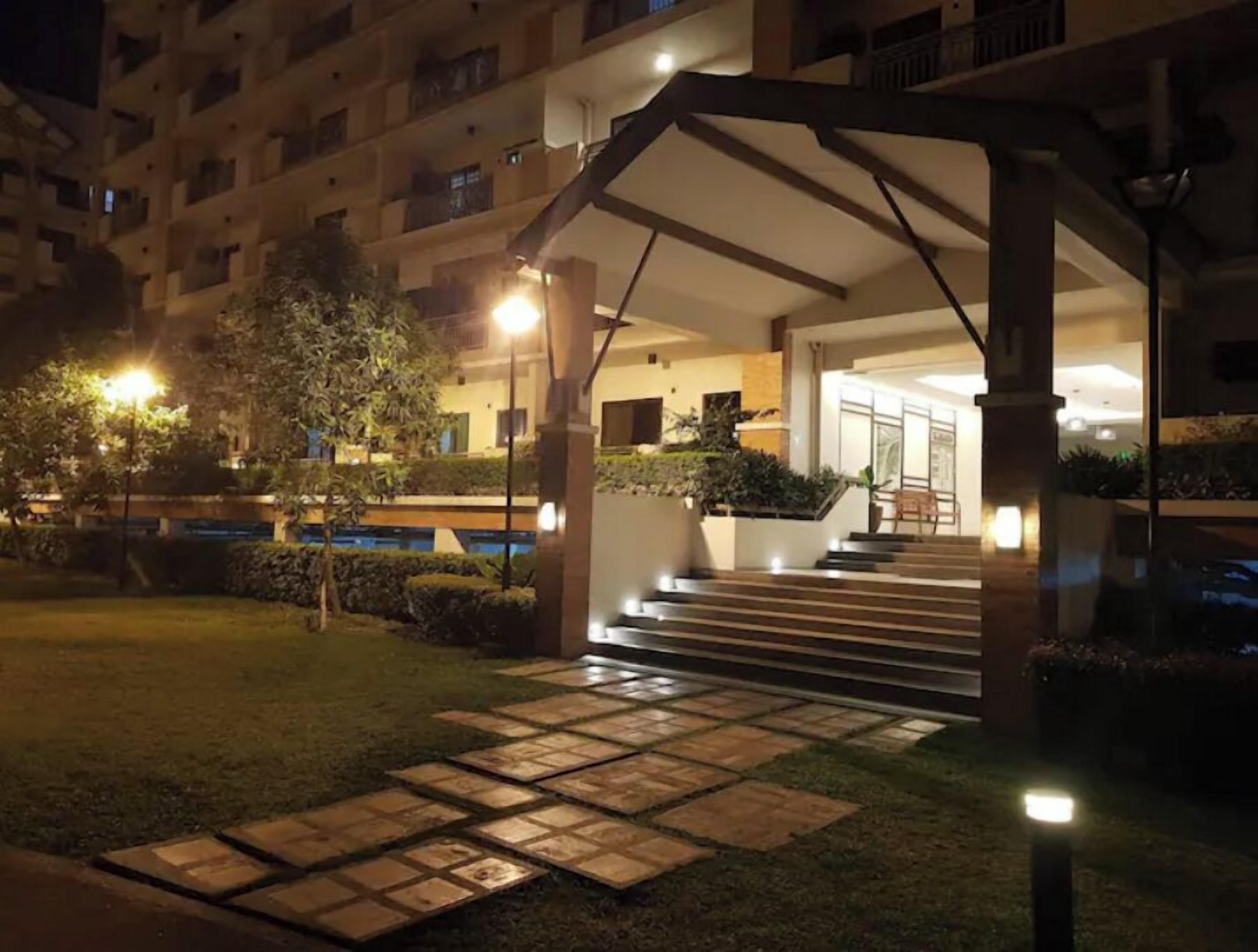 A well-lit outdoor area with a covered entrance, steps leading to a building, and a small garden with a tree and shrubs.