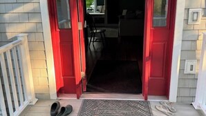 Entrada de casa com porta vermelha, varanda com tapete e um par de sapatos no chão.