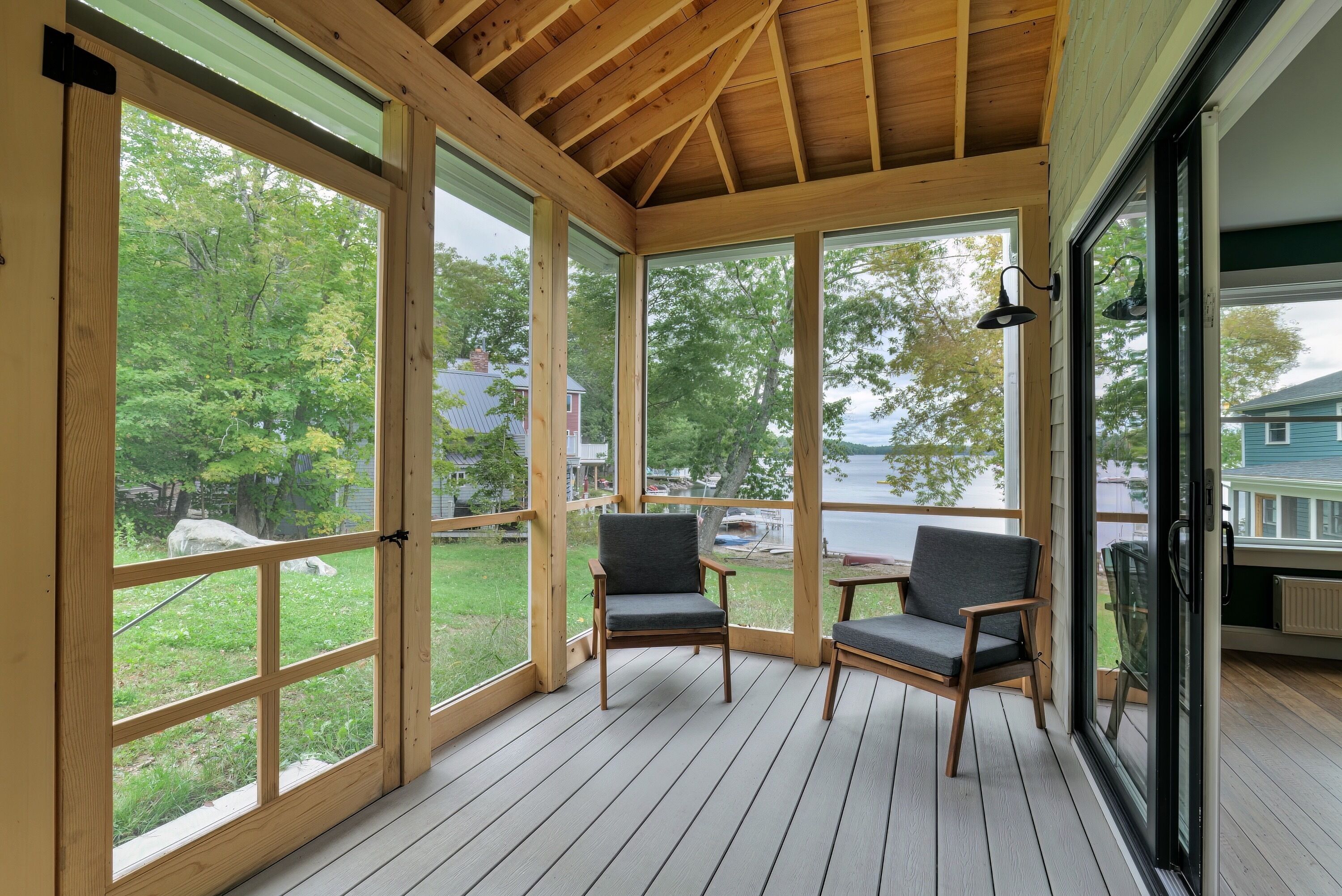 A covered porch with large windows, two chairs, and a view of a lake and trees.