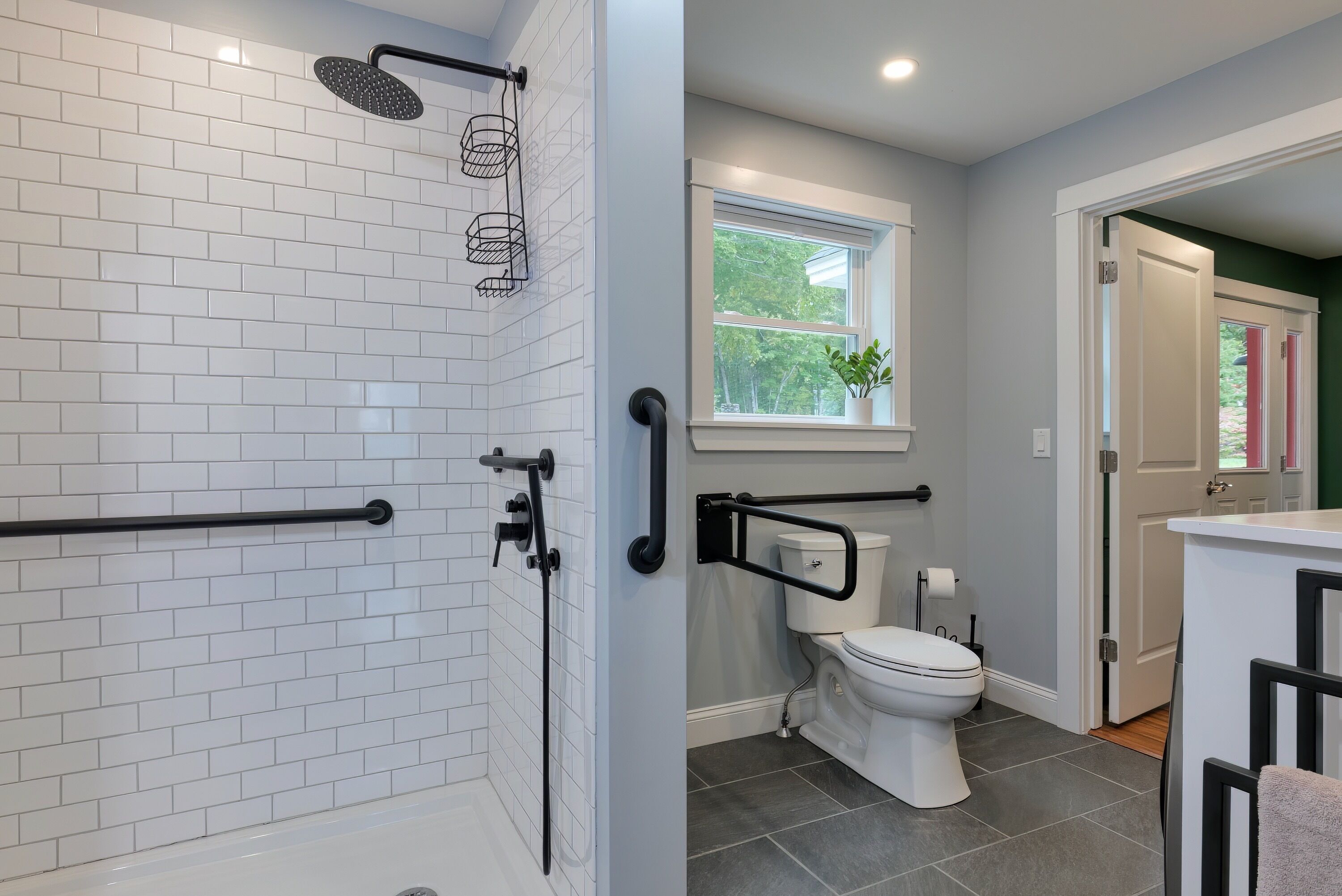 Une salle de bain moderne avec des carreaux de métro blancs, une douche équipée d’un pommeau de douche à main, des toilettes dotées d’une barre d’appui et une fenêtre donnant sur un espace verdoyant.