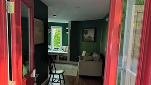 Uma porta que dá para um quarto com piso de madeira, uma cadeira, um sofá e uma janela com vista para o verde.