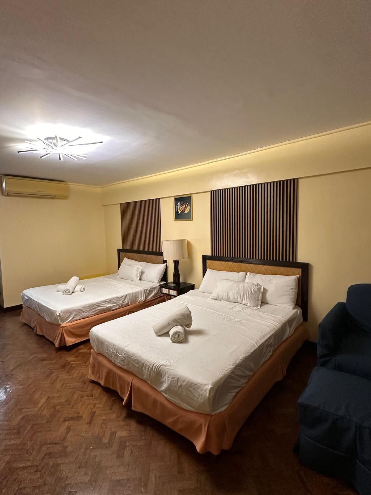 Quarto de hotel com duas camas, piso de madeira, luminária no teto e ar-condicionado fixado na parede.