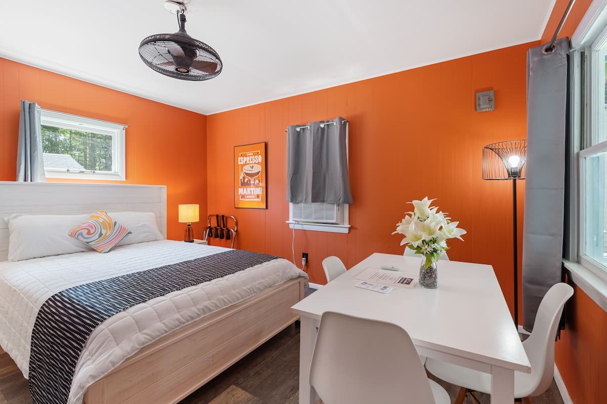 A hotel room with a bed, a desk, a chair, a fan, a window, and an air conditioning unit.
