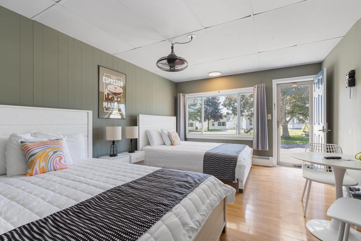 A hotel room with two beds, a desk, a chair, a window with curtains, and a ceiling fan.